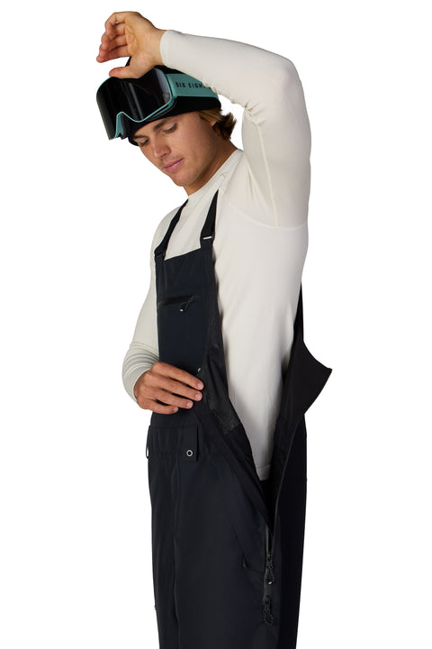 Man wearing black 686 shell bib pants with teal ski goggles and white base layer