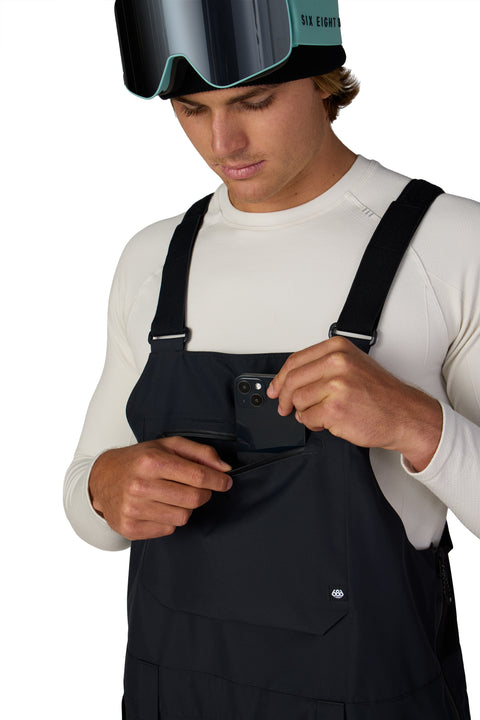 686 men's black shell bib pant with chest pocket, model inserting phone
