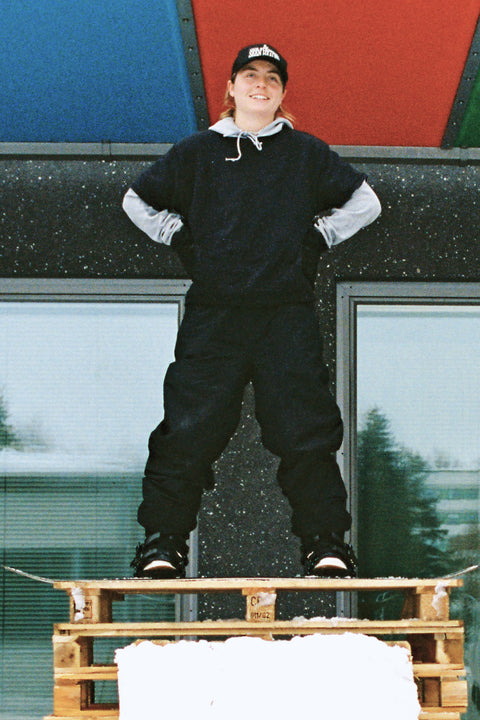 Woman standing confidently on wooden pallet wearing black shell snow pants and hoodie