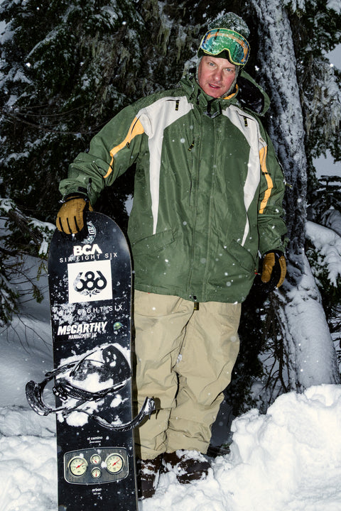 Man in green white and yellow 686 snowboard jacket holding board in snow