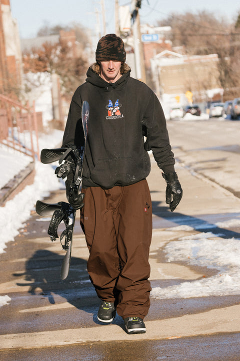 Man walking with snowboard wearing brown 686 Dojo Gore-Tex shell snow pants