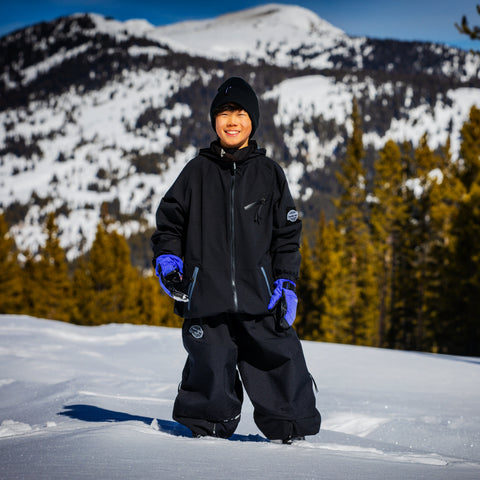 Child wearing full black shell jacket and snow pants on snowy mountain slope with evergreen trees