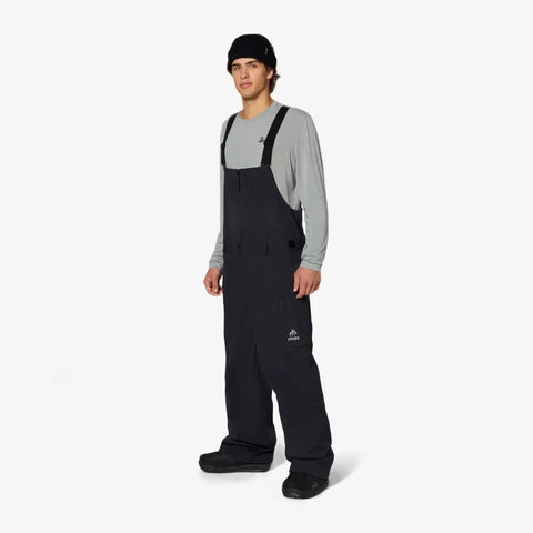 Male model wearing black Jones Snowboards bib snow pants with suspenders and gray base layer